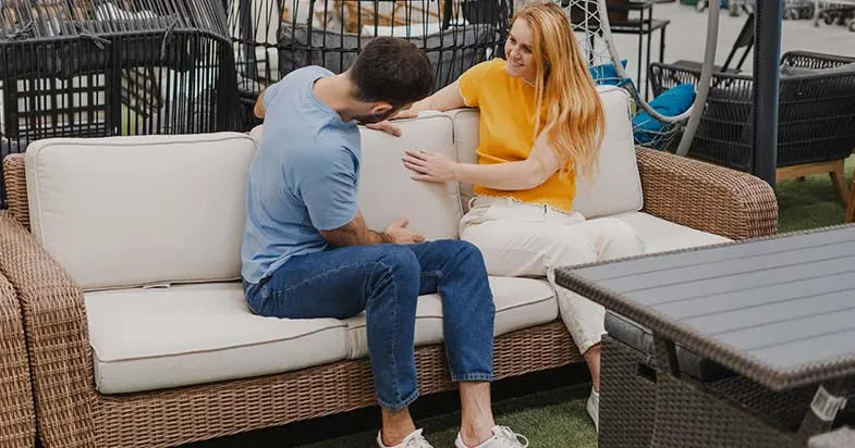 A couple sitting on a comfortable outdoor wicker sofa with cream-colored cushions, discussing furniture features in a showroom setting.