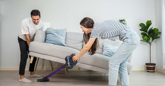 woman vacuuming fabric sofa while man lifts couch for deep cleaning in living room